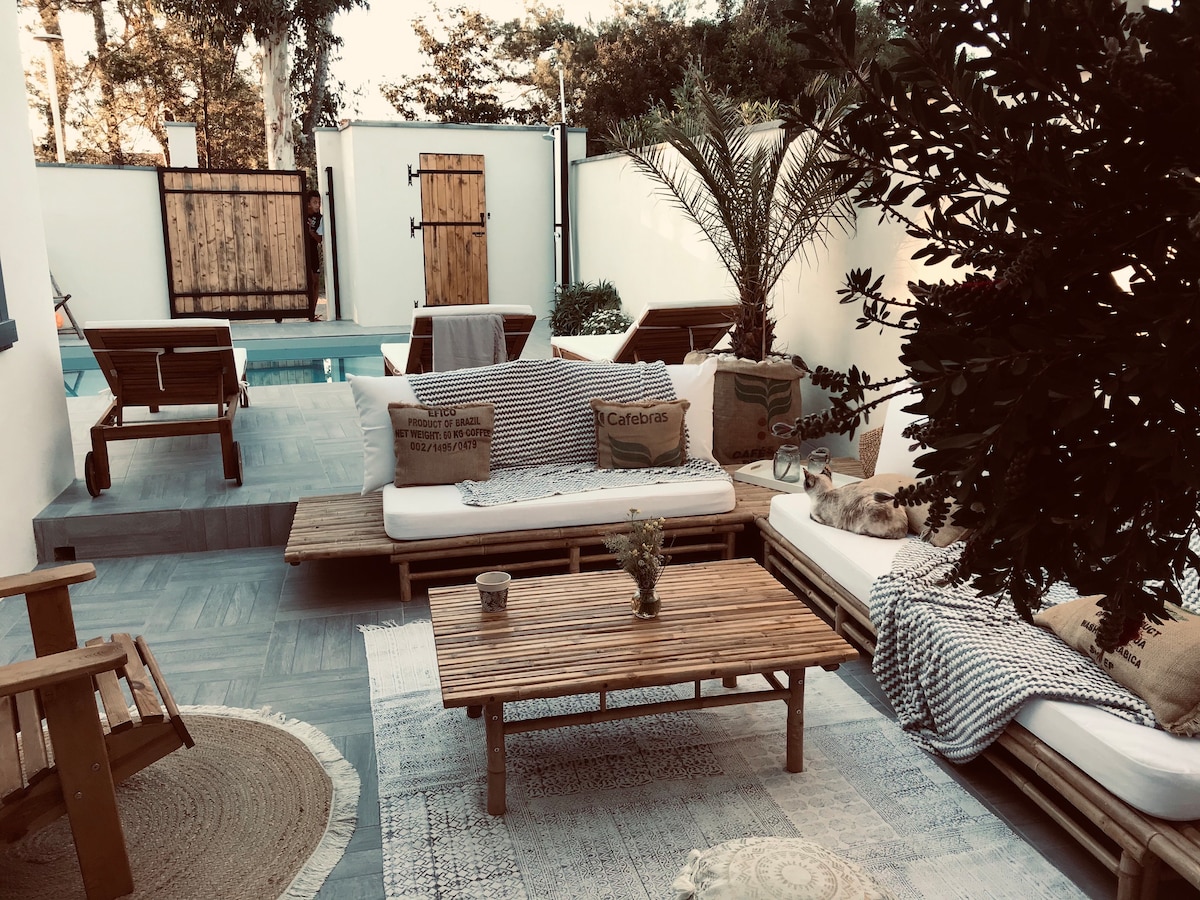 An outdoor seating area is featured with wooden lounge chairs and a low coffee table, surrounded by greenery. Soft cushions are placed on platforms, creating a relaxed atmosphere. The pool area is visible in the background, with a muted color palette enhancing the serene environment.