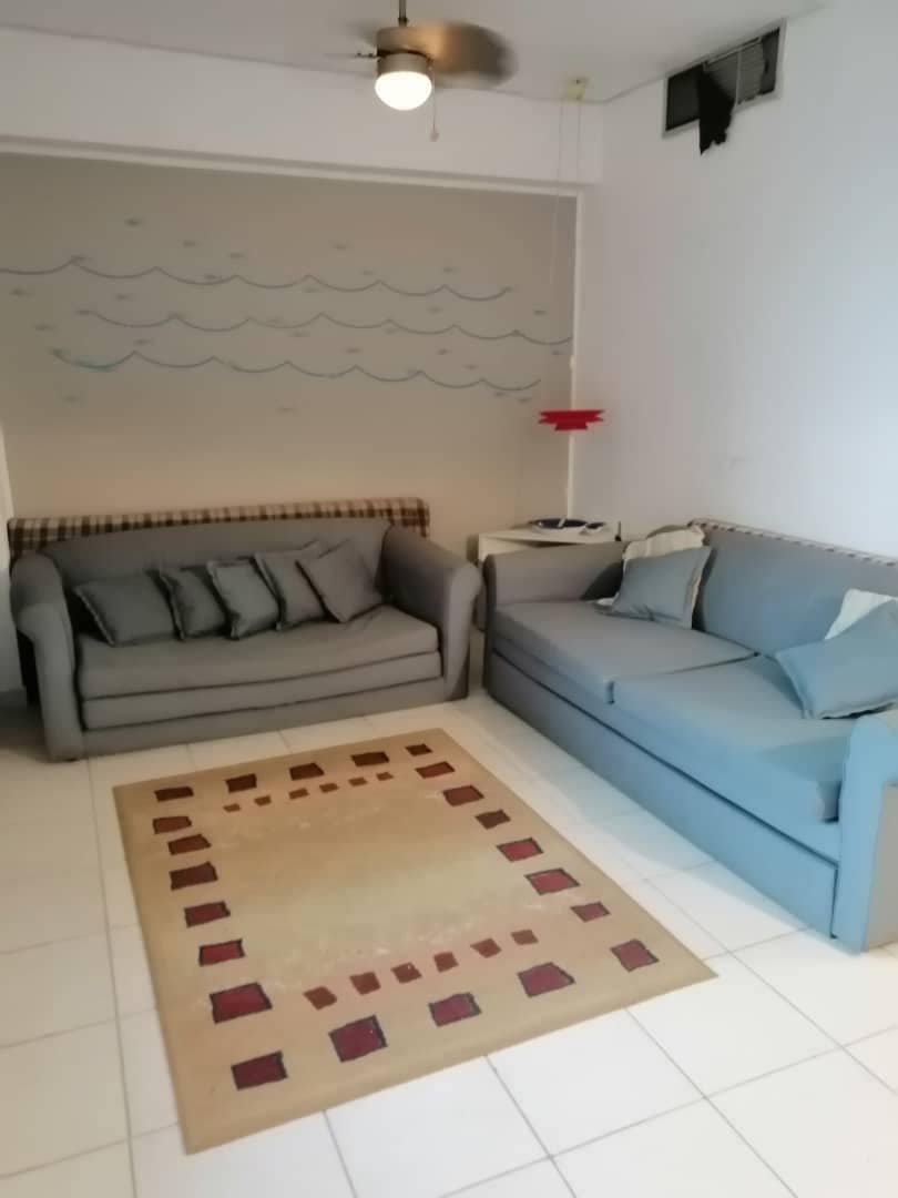 A bright living area features two upholstered sofas arranged around a patterned rug. A light fixture hangs from the ceiling, and a wall mural depicts gentle waves. Light-colored tiles enhance the spacious feel of the room.