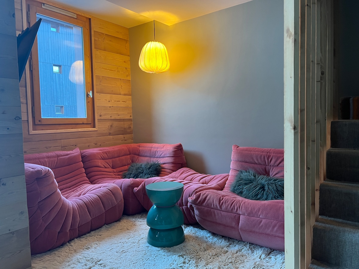 A seating area features soft, curved furniture in a warm pink hue, complemented by a turquoise side table. Soft textures are present in the room, highlighted by natural wood paneling and a warm light fixture, enhancing the cozy atmosphere.