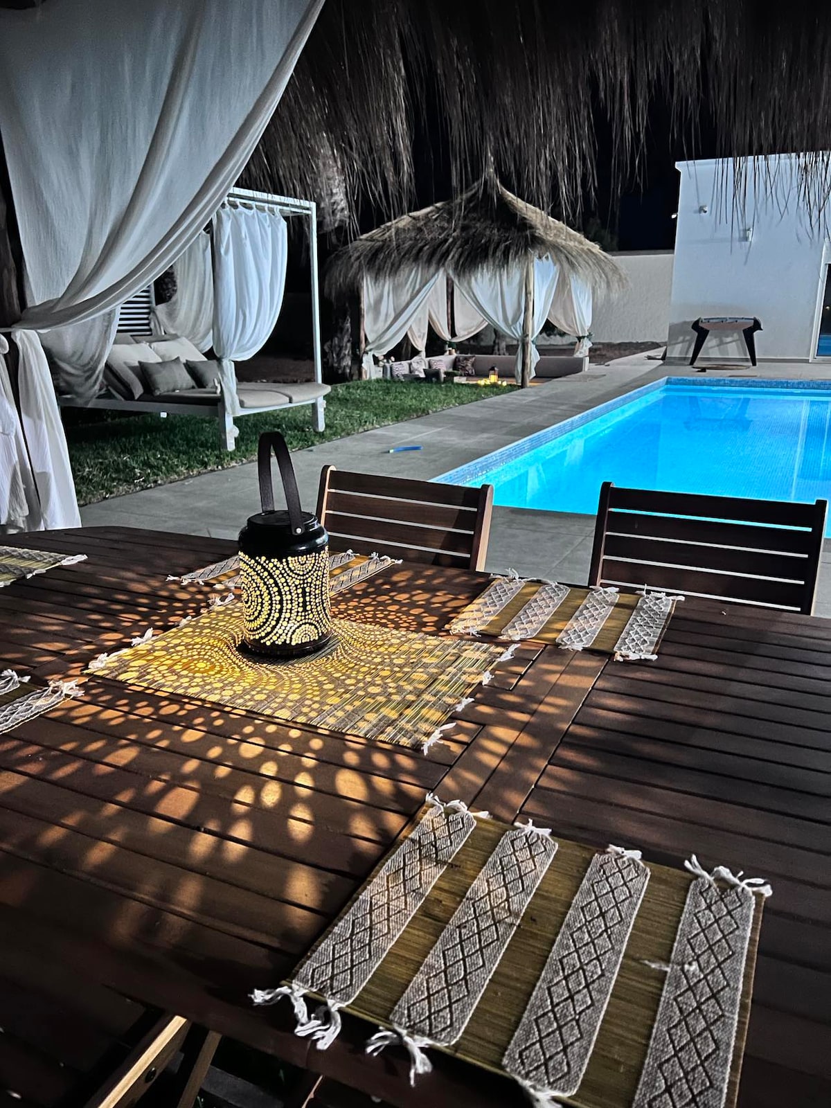 A wooden dining table is set with woven placemats, illuminated by a decorative lantern. The background features a serene pool, framed by lounging areas with elegant canopies, all under soft, ambient lighting enhancing the evening atmosphere.