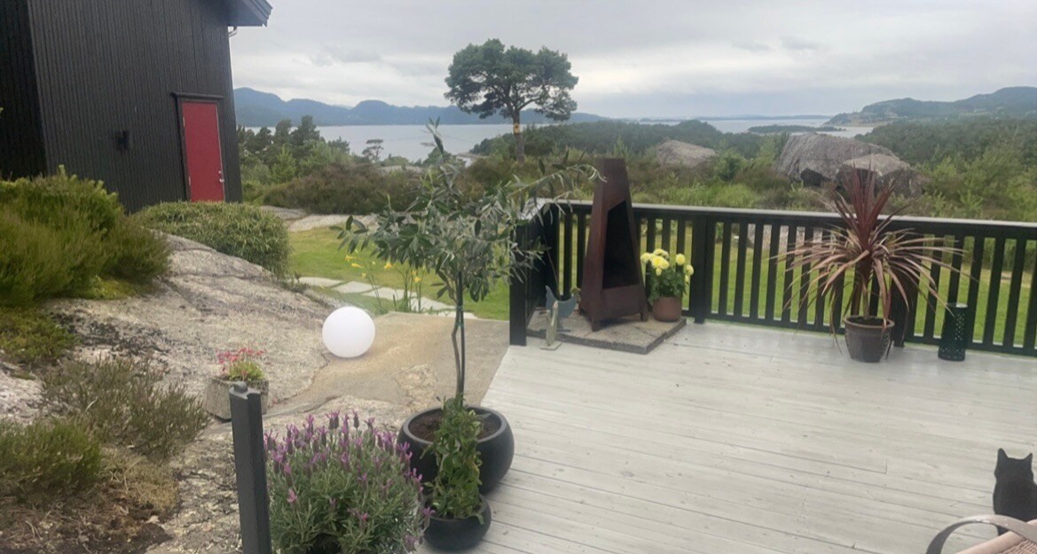 A spacious terrace is surrounded by greenery and rocks, featuring potted plants and decorative items. A view of the fjord and hills is visible in the background, while a sleek fire feature adds a modern touch. Soft, natural light fills the outdoor space.