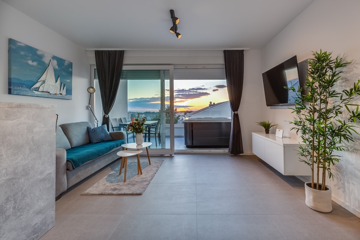 A modern living area features a comfortable sofa, a low coffee table, and a wall-mounted TV. Large sliding doors offer access to a terrace with a hot tub and sunset views. Soft lighting complements the relaxing ambiance, while potted plants add a touch of greenery.