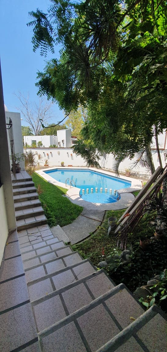 A serene view of a private pool area is shown, surrounded by lush greenery and softly swaying trees. Clear blue water reflects the sky, while a stone pathway leads gently down from the upper level. The space is designed for relaxation and tranquility.