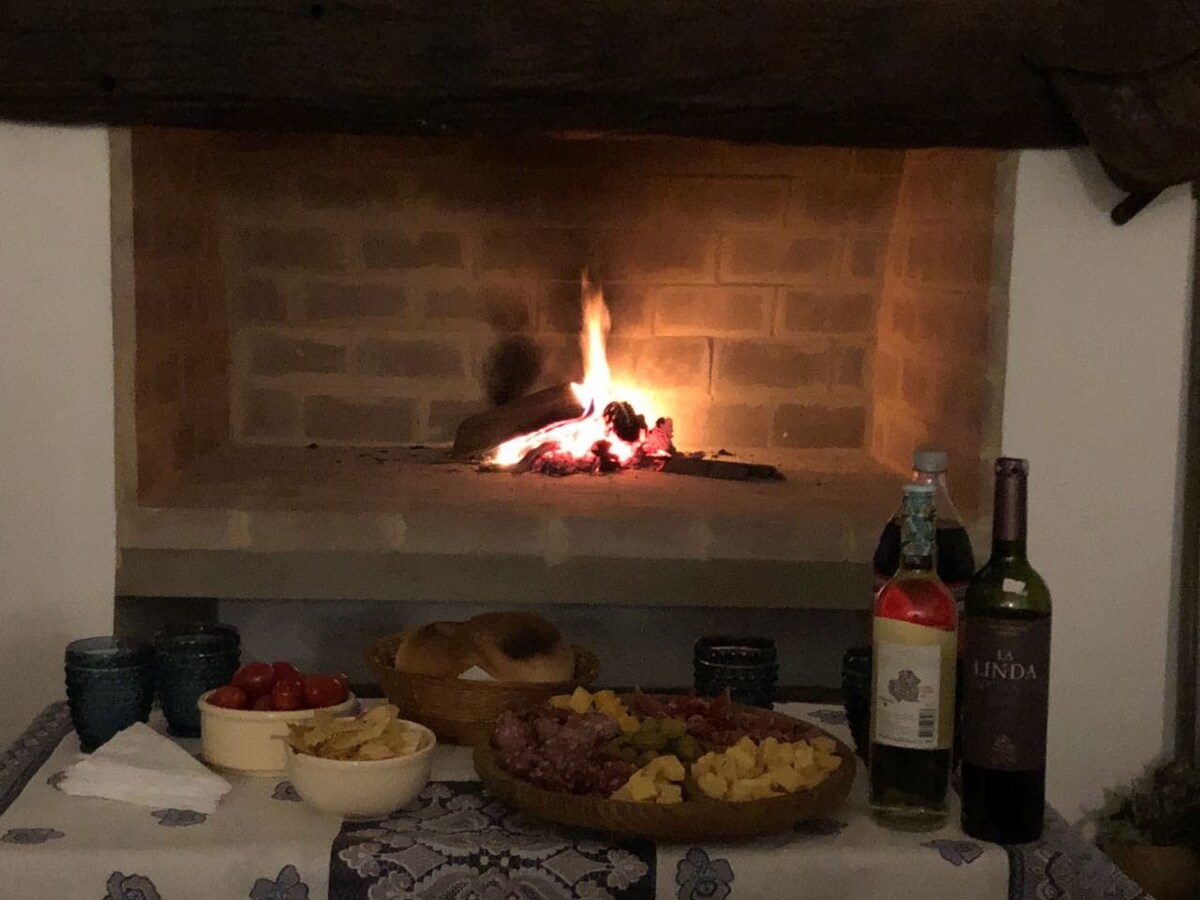 A rustic fireplace is visible, with flames gently flickering against the brick backdrop. A table is set nearby, featuring an assortment of cheeses, cured meats, bread, and fresh produce, accompanied by bottles of wine and drinks. Blue ceramic dishes complement the warm atmosphere.