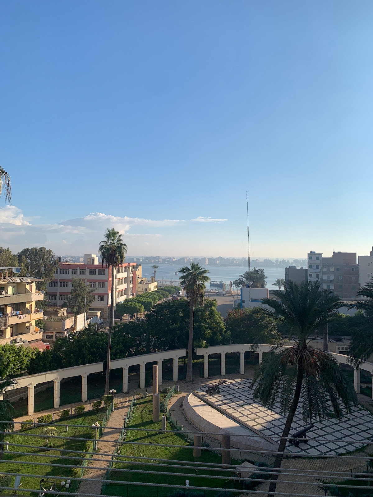 A scenic view reveals a mix of residential buildings and lush greenery, with a large circular stone patio in the foreground. Palm trees are visible, and a stretch of water can be seen in the background under a clear blue sky.