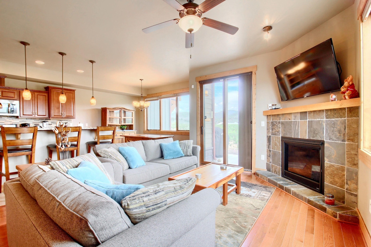 A spacious living area features comfortable gray sofas adorned with blue pillows, positioned around a wooden coffee table. A flat-screen television is mounted above a stone fireplace, while large windows allow natural light and mountain views to fill the space.