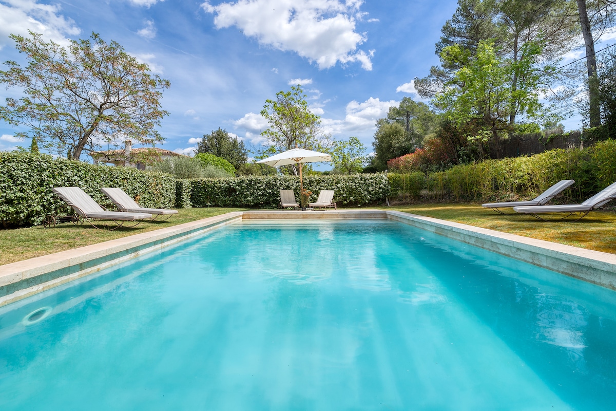A spacious outdoor pool is surrounded by lush greenery and features comfortable loungers for relaxation. A large umbrella provides shaded seating near the water's edge, while a clear blue sky enhances the tranquil ambiance.