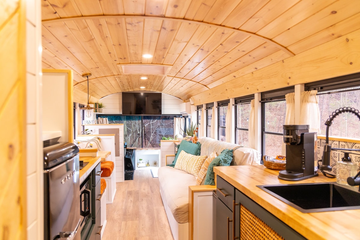 The interior of the bus features a warm wood ceiling and spacious layout. A cozy seating area is created with a light-colored sofa adorned with colorful throw pillows. Natural light flows through large windows, illuminating the kitchen area equipped with modern appliances and a coffee maker.