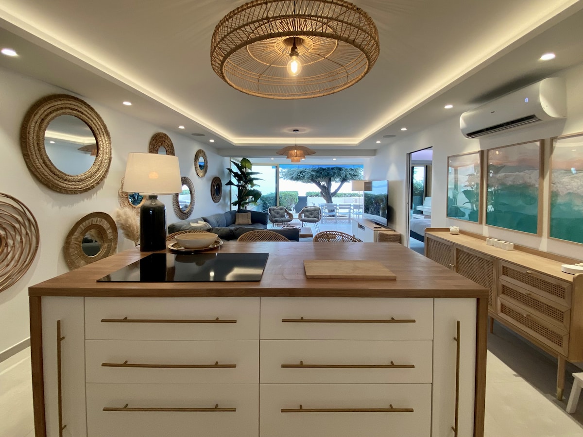 A modern kitchen island with a wooden countertop serves as the centerpiece in an open living area. Decorated with round mirrors and warm lighting, the space features a seamless transition to a terrace with sea views, highlighting a relaxed coastal ambiance.