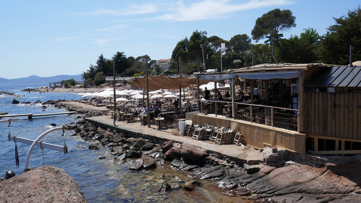 A seaside restaurant features wooden seating and a shaded area overlooking the water. Smooth, red rock formations line the shore, with gentle waves lapping at the edges. Nautical elements such as masts and small boats can be viewed in the distance.
