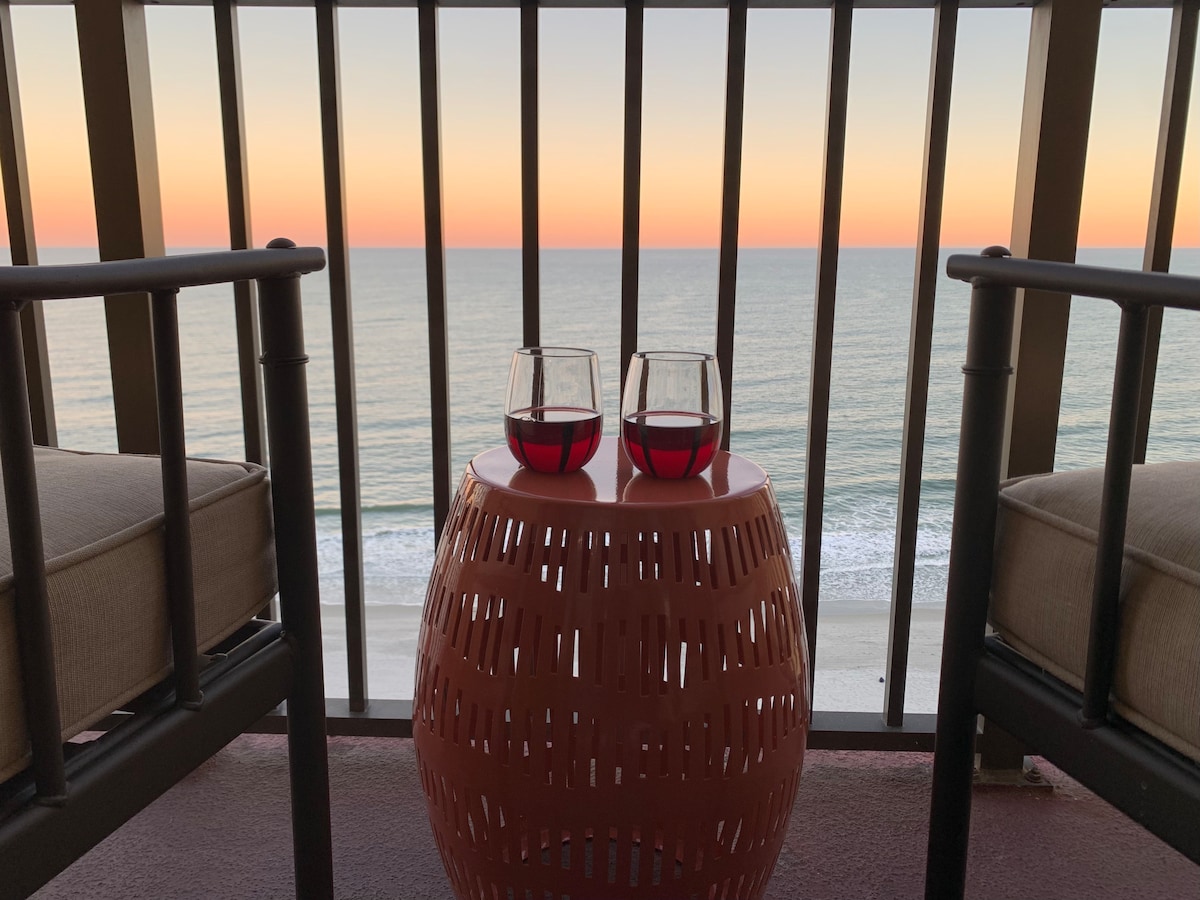 A private balcony is presented with two glasses of red wine resting on a round, decorative table. Comfortable seating is positioned on either side, offering unobstructed views of the ocean and a colorful sunset illuminating the sky.