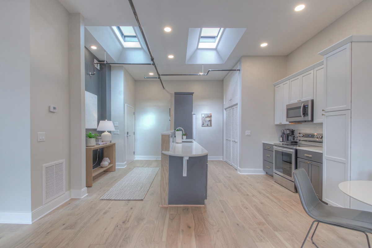 An airy kitchen is featured, showcasing modern white cabinetry and sleek stainless steel appliances. Skylights allow natural light to pour in, highlighting the spacious countertops. A central island provides additional workspace, complemented by a dining chair positioned nearby.