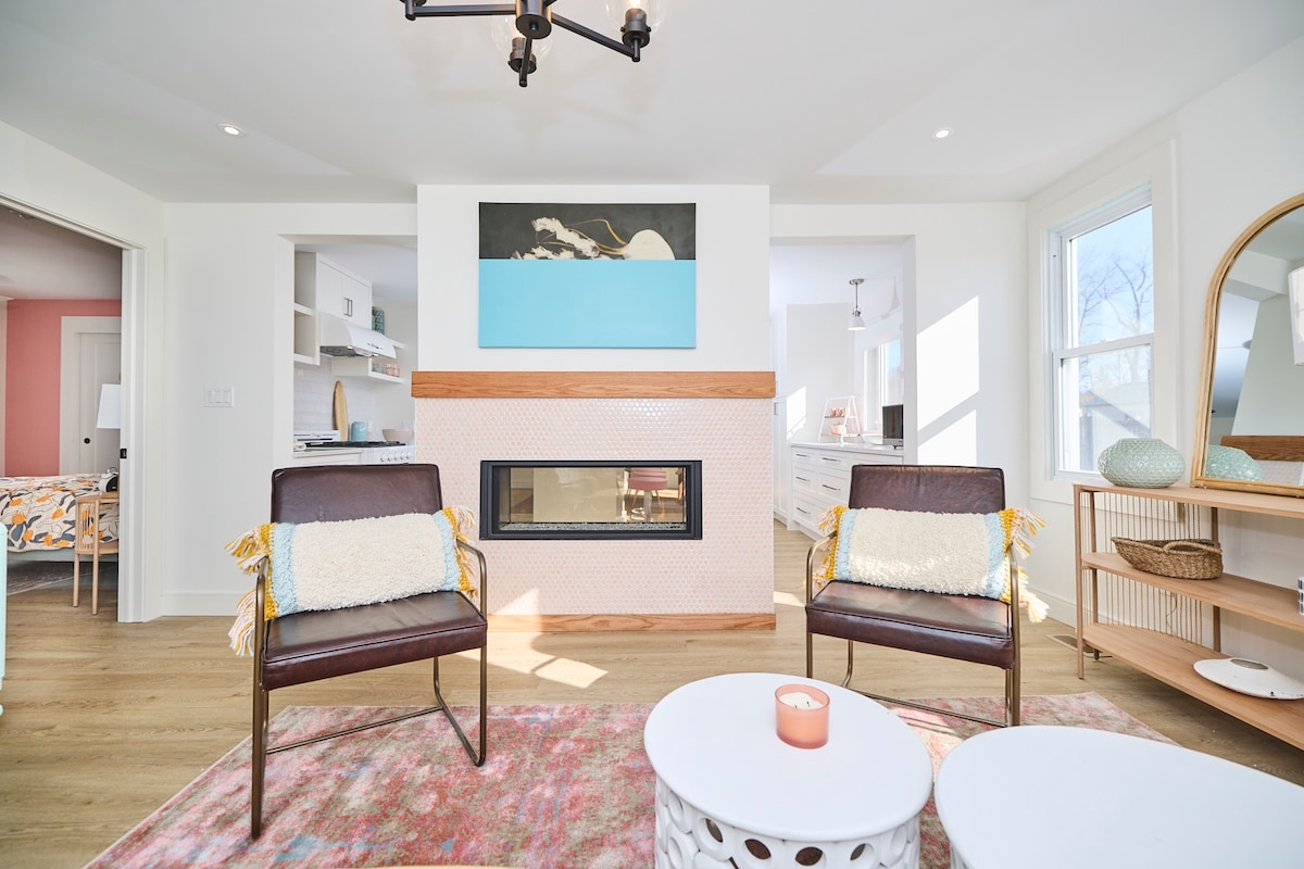 A contemporary living area is displayed, featuring two stylish chairs positioned beside a round table. A gas fireplace is centrally located, complemented by a colorful piece of art above it. Natural light fills the space through large windows, enhancing the welcoming ambiance.