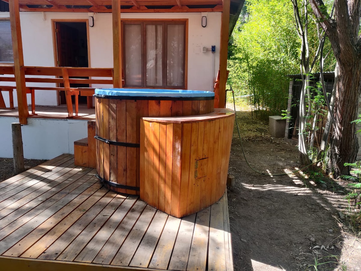 A wooden hot tub is set on a spacious wooden deck, surrounded by greenery. The tub's exterior features natural wood tones, and a small built-in area is present for seating. Nearby, a cabin is partially visible, with large windows and a welcoming entrance.