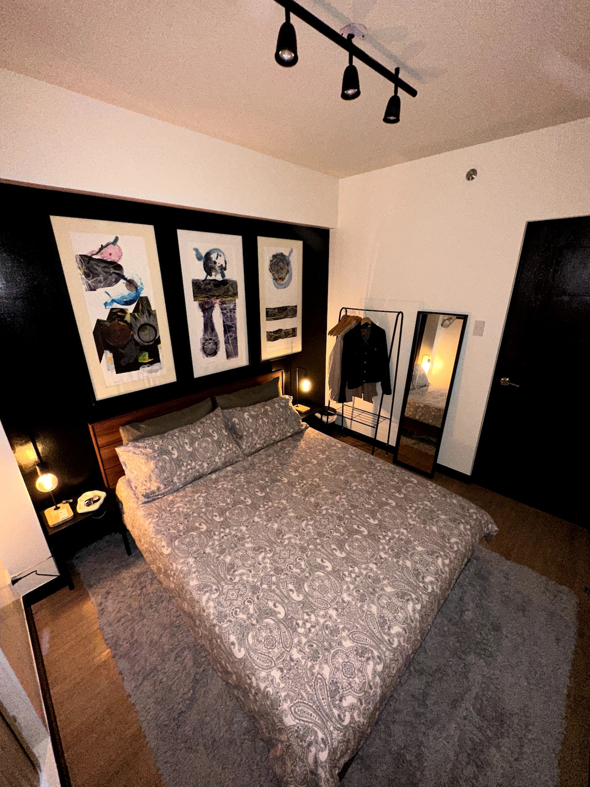 A cozy bedroom features a queen-size bed with a patterned duvet and two decorative pillows. Artwork is displayed on the wall behind the bed, complemented by stylish bedside lamps. A full-length mirror and a clothing rack are visible, adding functionality to the space.