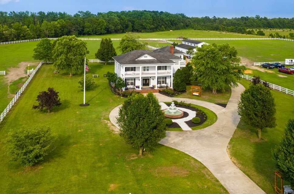 A spacious estate is set within a lush landscape, featuring manicured lawns and mature trees. A circular driveway leads to the entrance, which has a central fountain and is framed by neatly arranged bushes. Outbuildings and open fields are visible in the background.