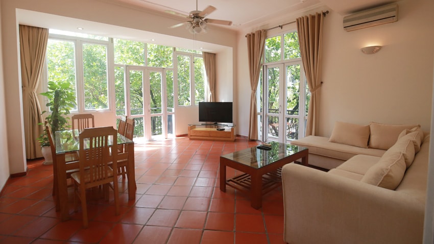 A spacious living area is filled with natural light, featuring large windows that invite outdoor views. A comfortable sofa is positioned beside a wooden coffee table. A dining set occupies one corner, while a television sits on a media console against the wall.