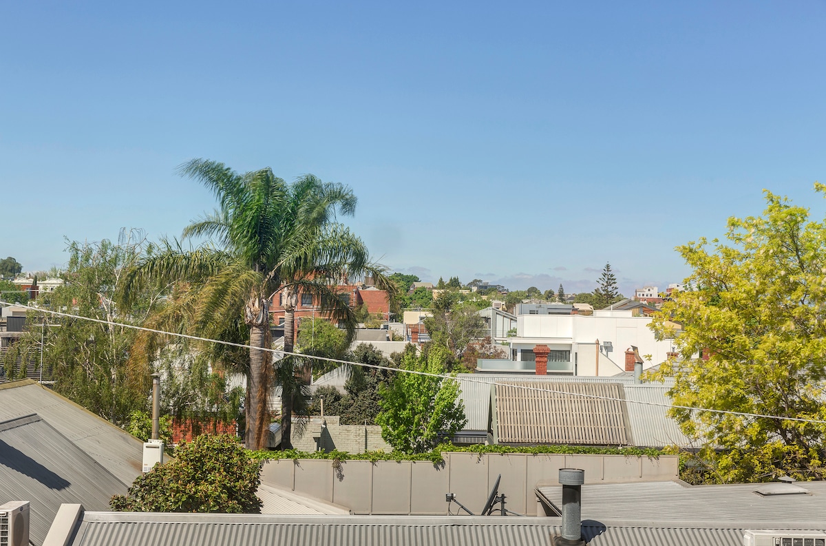 A view from above showcases a variety of rooftops and greenery, including palm and leafy trees, framed against a clear blue sky. The scene reflects a vibrant neighborhood, with hints of architecture and natural elements visible in the distance.