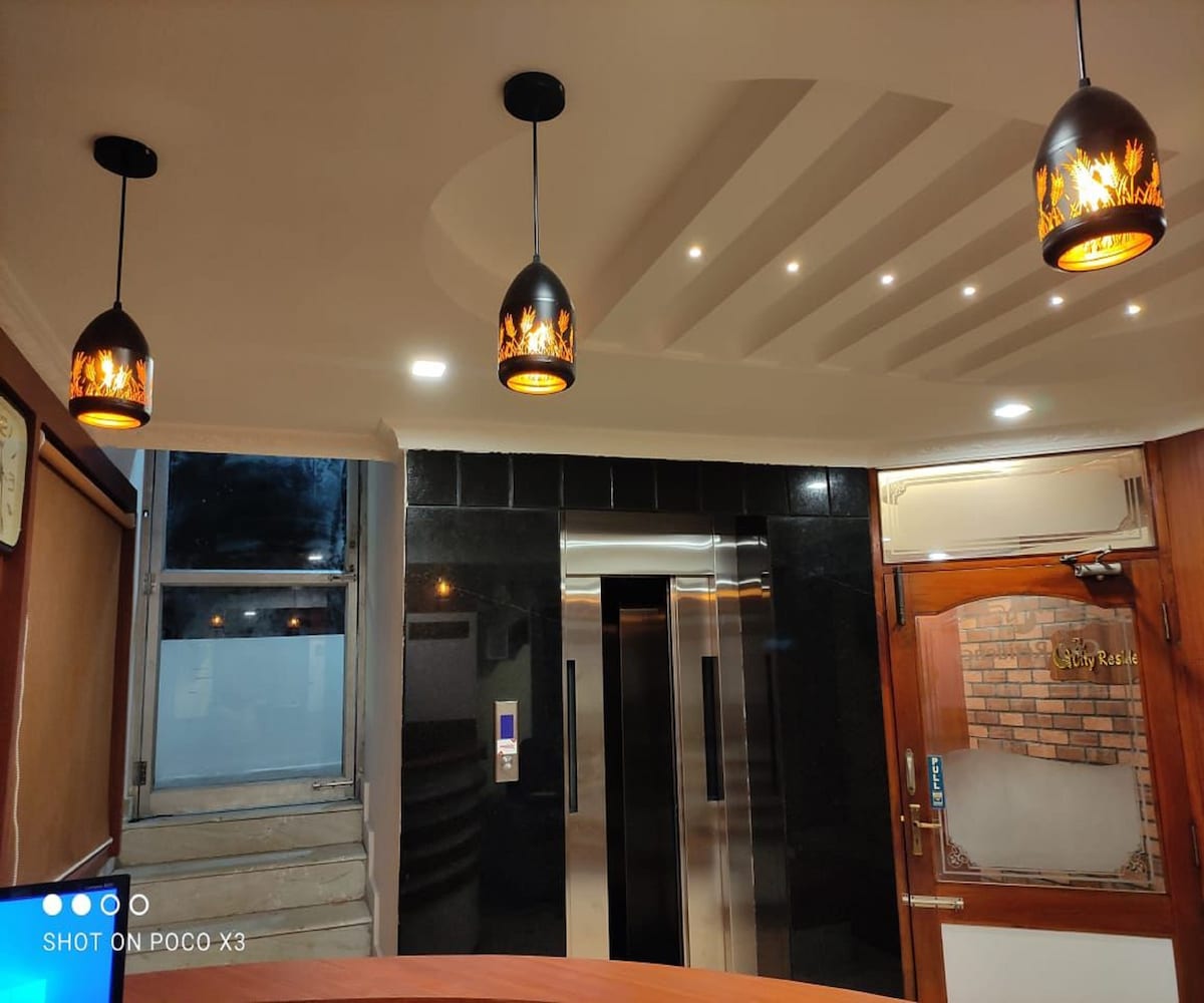 The entrance area features modern pendant lights with a warm glow. An elevator is positioned against a sleek black wall, while large windows allow natural light. A wooden reception desk is visible, creating an inviting transition from the outside.