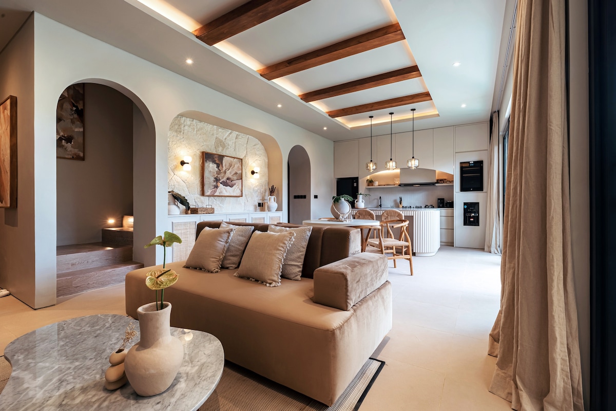 An open-concept living space is showcased, integrating a fully equipped kitchen and dining area. The stylish furniture includes a soft sofa and a marble coffee table, with warm ambient lighting accentuating the ceiling beams. Large glass doors invite ample natural light.