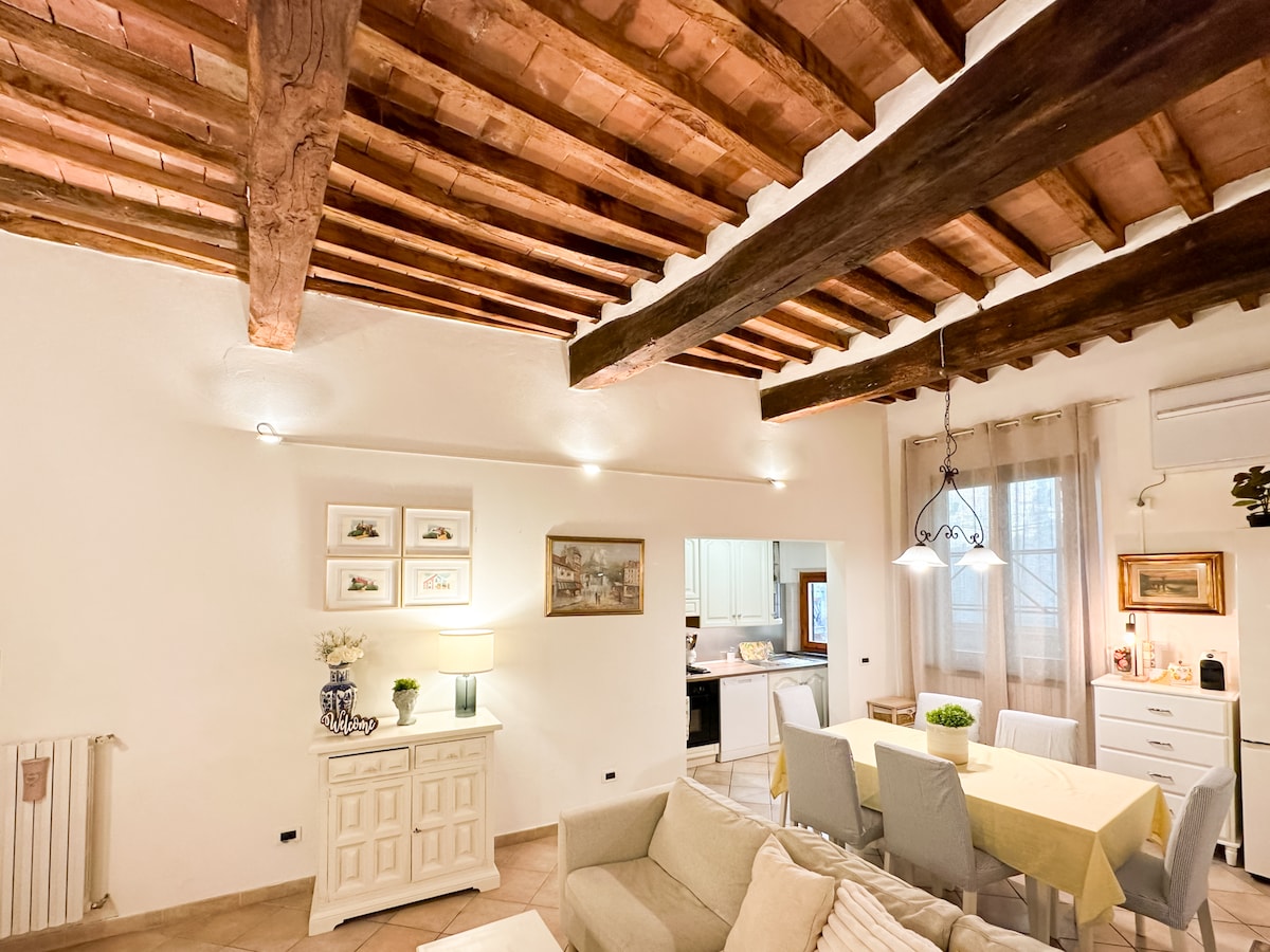 An inviting living area is highlighted by exposed wooden beams and soft lighting. The space features a comfortable sofa and a light-colored table set for dining. A vintage cabinet adds character, while a doorway leads to a fully equipped kitchen, enhancing the cozy ambiance.