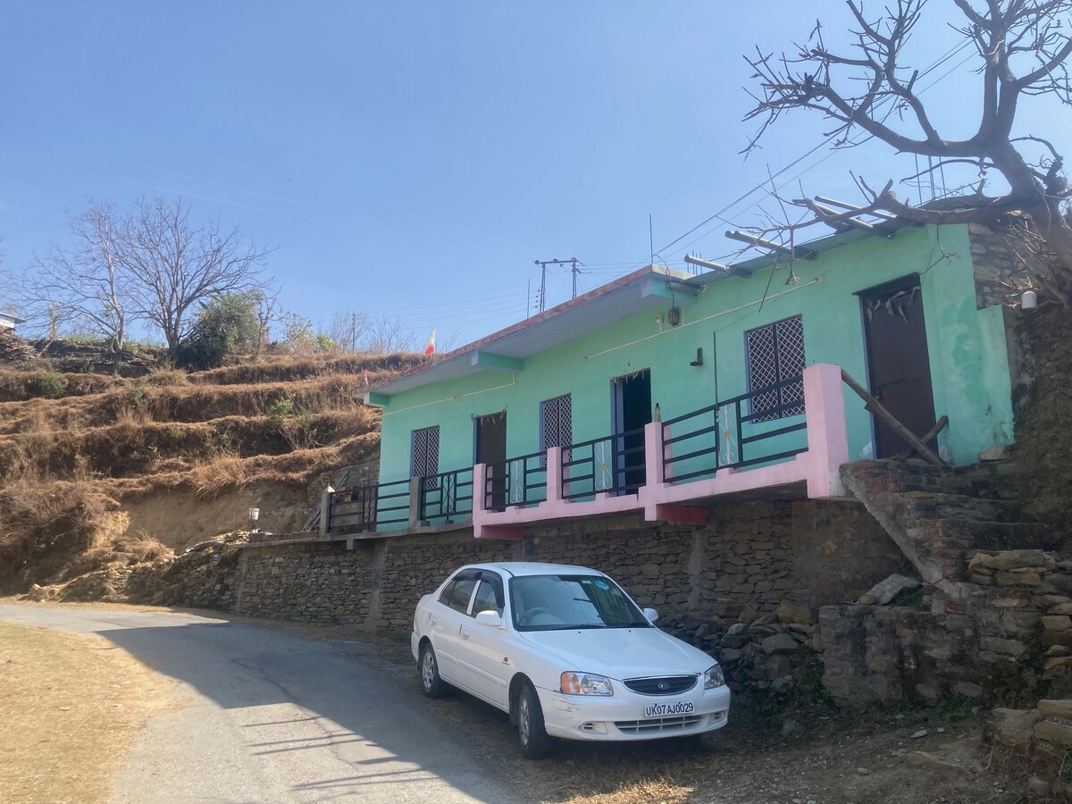 A modest single-story home is set against a hillside, featuring a light green exterior with pink accents. A car is parked in front of the house, and a pathway leads to the entrance, bordered by grassy areas. Bare trees are visible in the background.