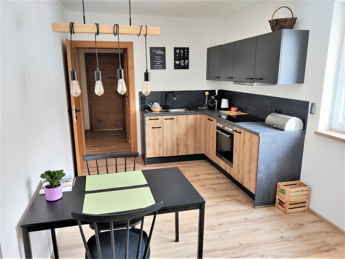 A modern kitchen is displayed, featuring wooden cabinetry and sleek black countertops. A dining table with a single black chair is positioned under a pendant light with visible bulbs. An entryway can be seen in the background, adding depth to the space.