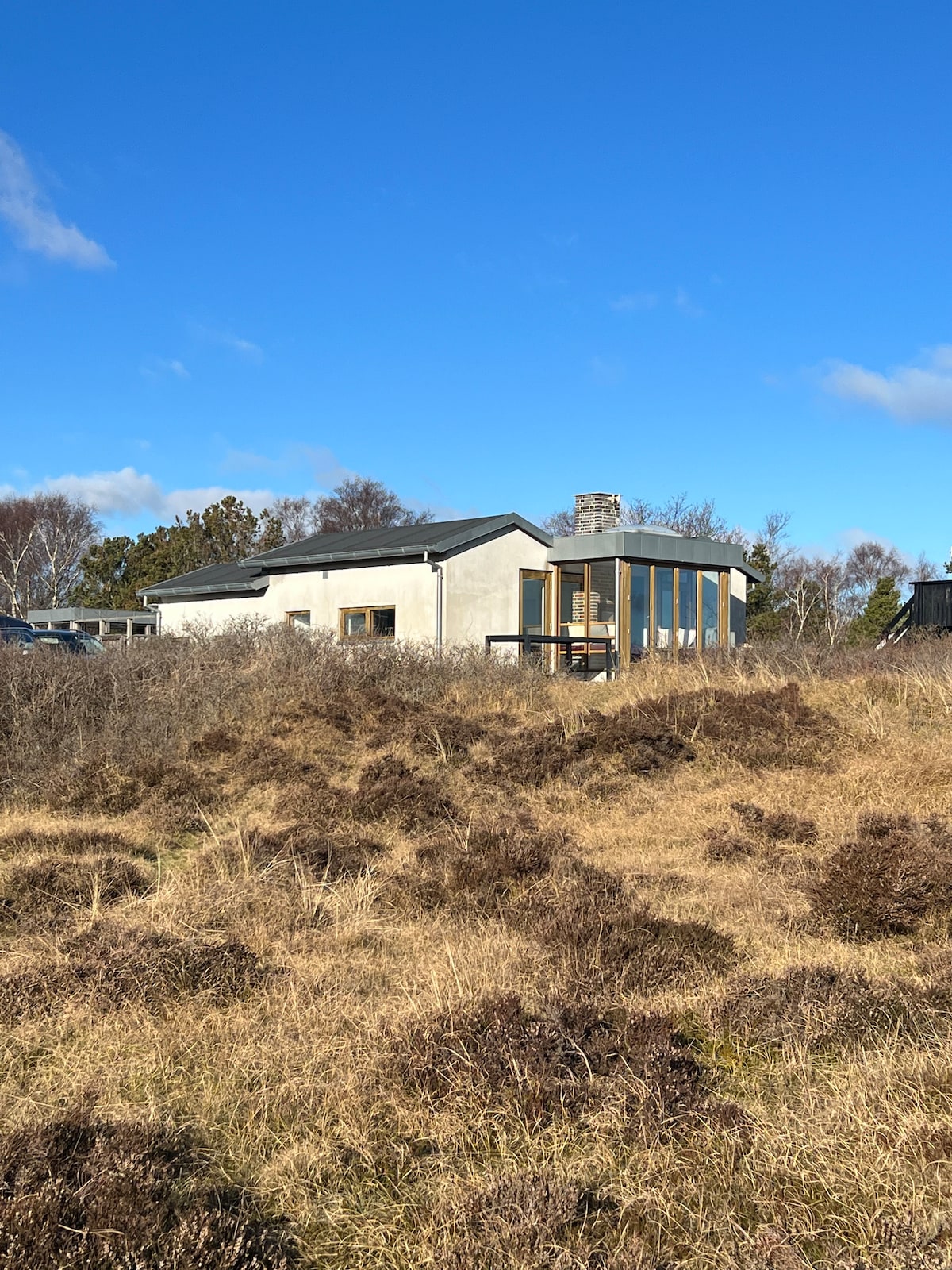 A modern summer house is depicted, featuring a combination of smooth facades and large glass elements. The building is set against a backdrop of natural landscape, with low vegetation and open skies, creating an inviting appearance in a tranquil outdoor setting.