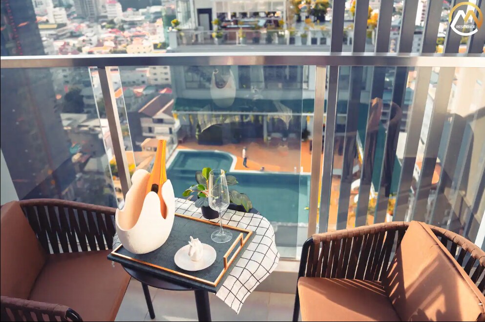 A private balcony is equipped with two wicker chairs and a small table. A glass of beverage and a small vase with greenery rest on the table, overlooking the city and the infinity pool below.