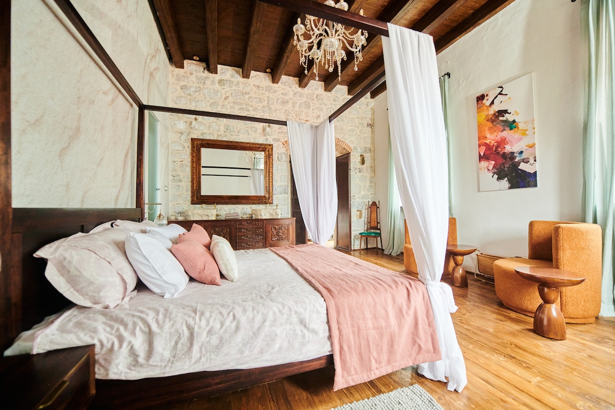 A spacious bedroom features a large wooden bed draped with flowing white fabric. The floor is made of warm wood, and natural light filters through light green drapes. Elegant artwork adorns the wall, and two soft armchairs provide a cozy seating area.