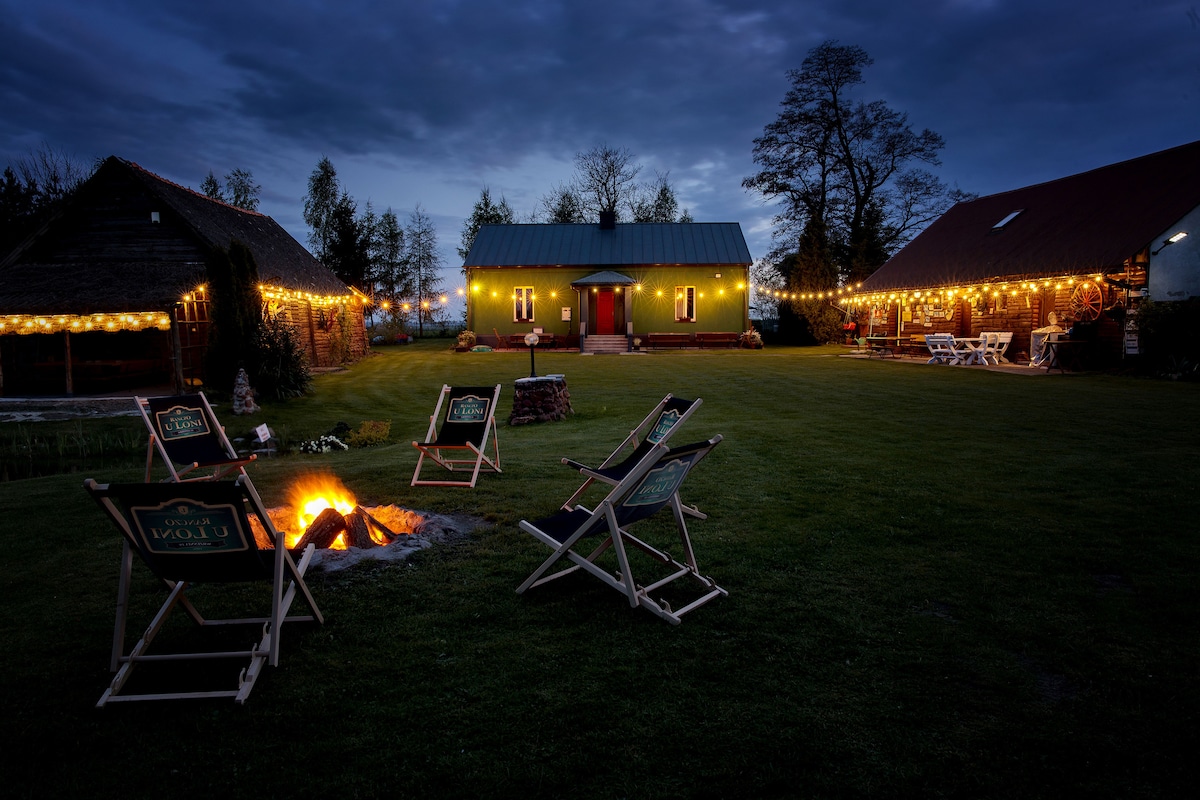 A spacious outdoor area is illuminated by string lights attached to nearby structures. Several chairs are arranged around a central fire pit, creating a cozy gathering space. The sky is filled with evening hues, enhancing the serene ambiance of the retreat.