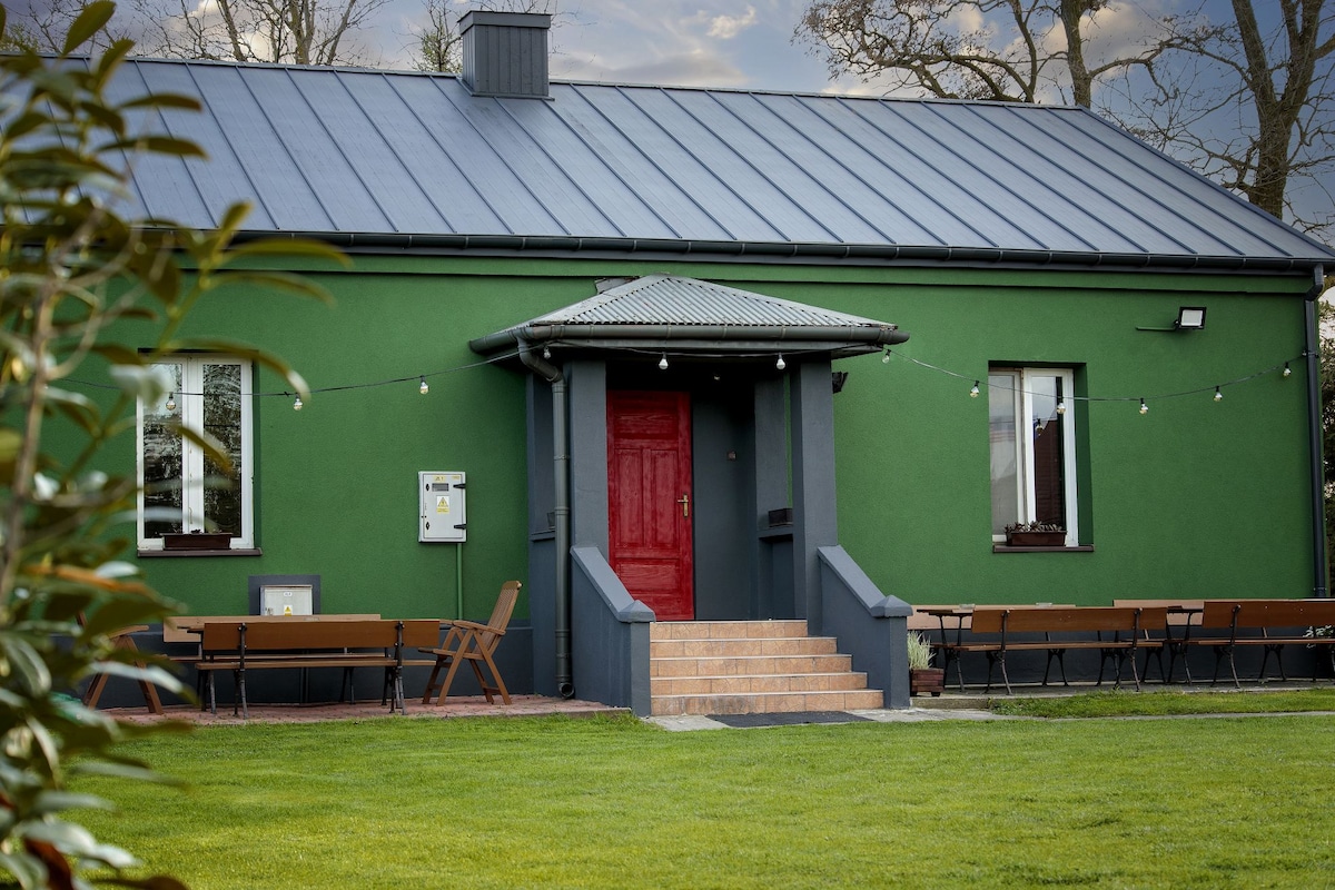 A vibrant green exterior of a house is complemented by a striking red front door. The entryway features steps leading up to the door, with outdoor seating arranged on either side. String lights add a touch of character to the welcoming ambiance.