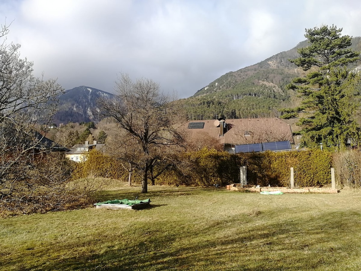 An expansive garden area is visible, featuring a grassy landscape with patches of sunlight. In the background, mountains rise against a cloudy sky, while a residential building with solar panels is nestled among trees and hedges. The garden offers ample open space for relaxation.