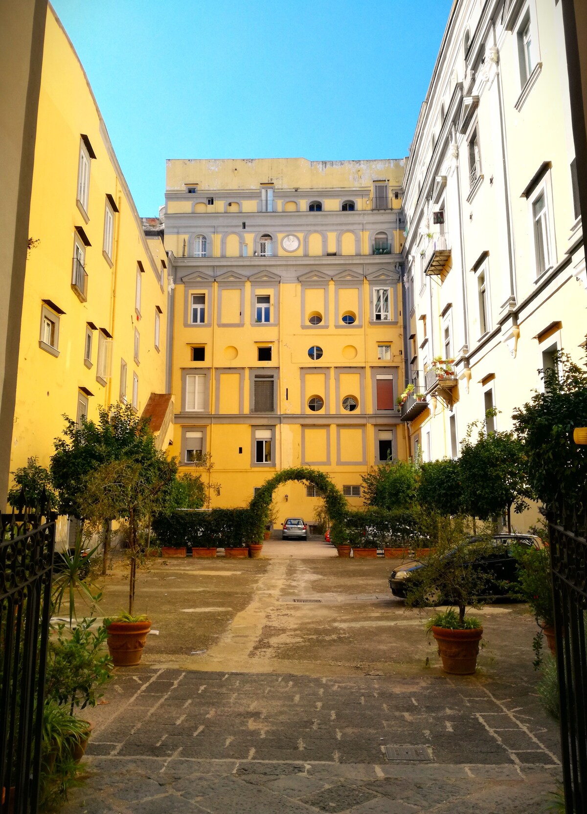 An open courtyard is surrounded by yellow and grey buildings, with trees in terra-cotta pots lining the pathway. The area is highlighted by clear blue skies, and a vehicle is parked at the end of the courtyard, creating a sense of calm and accessibility.