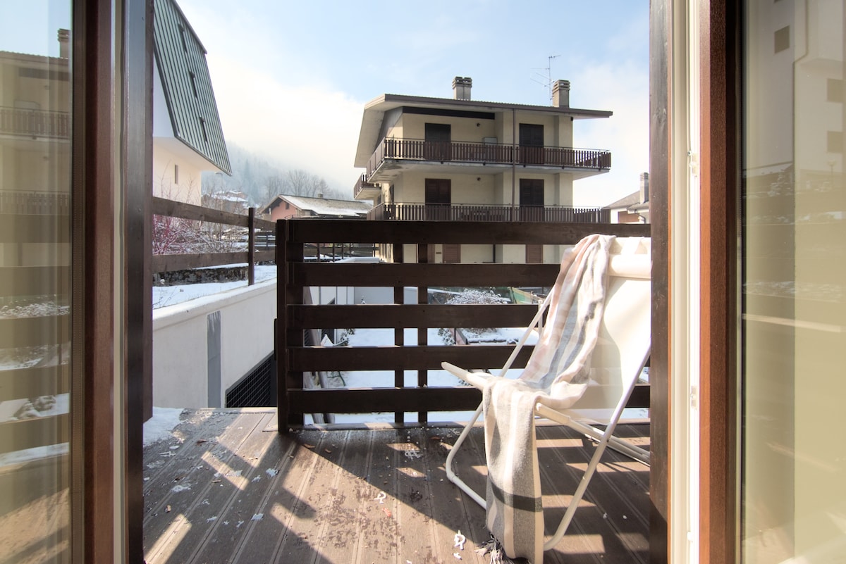 A small balcony is depicted, featuring a comfortable lounge chair and a compact wooden railing. Snow-covered surroundings create a serene atmosphere, with a view of neighboring buildings and mountain peaks in the distance. Soft sunlight filters in, adding warmth to the outdoor space.