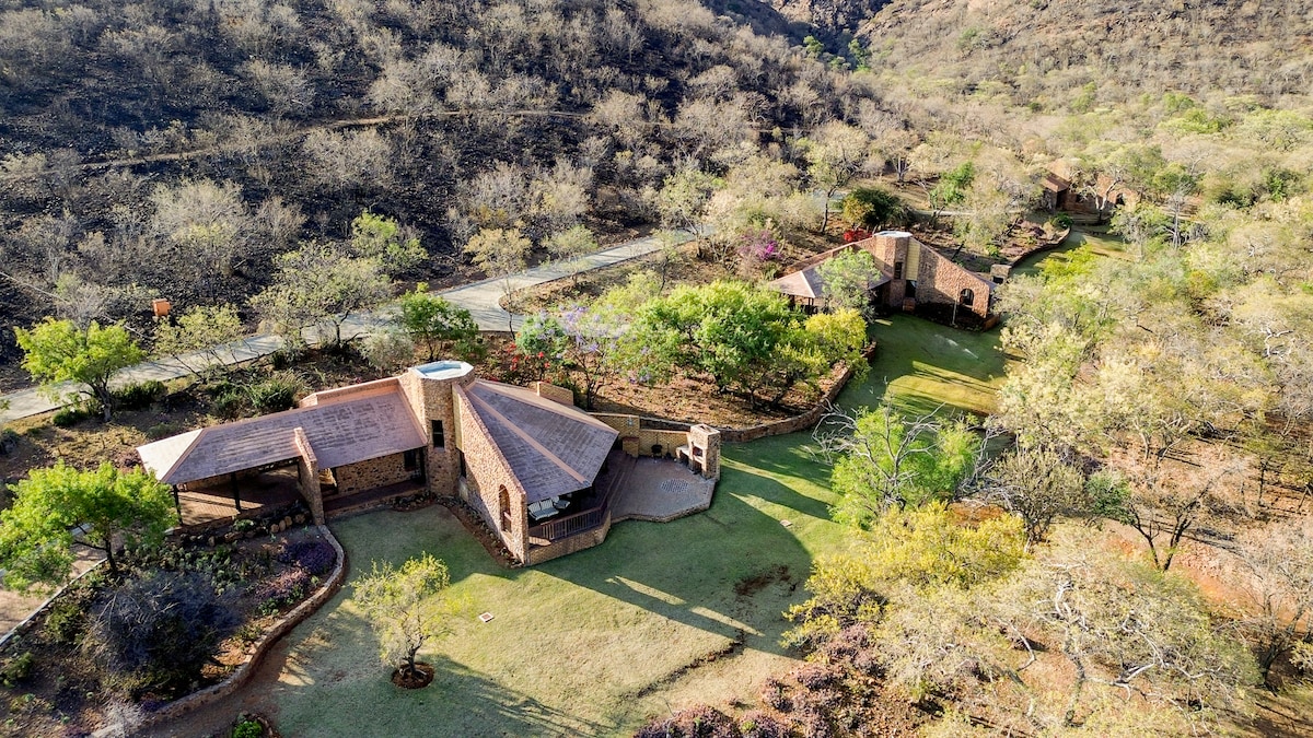 An aerial view of Leopards Cove showcases multiple cabins nestled among greenery in a natural landscape. The property features areas of manicured lawns, tree-lined paths, and surrounding hills, highlighting its serene and peaceful setting.