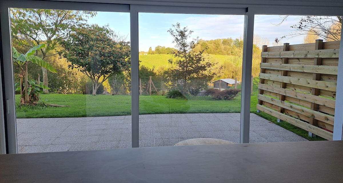 A view from the indoors shows expansive greenery outside, with a mix of trees and plants visible. A well-maintained lawn stretches out towards a distant shed, and a wooden fence lines one side. The scene is framed by large glass doors, inviting natural light.