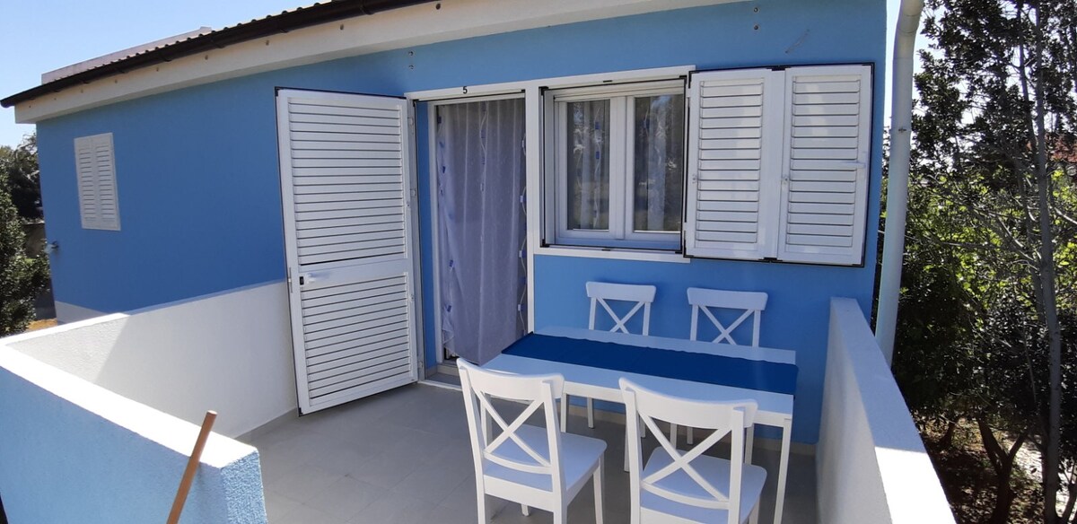 A terrace is adorned with a blue table surrounded by four white chairs, set against a backdrop of blue walls. Two windows with white shutters open to the outside, showcasing the garden views. The floor is tiled, adding to the functionality of the space.