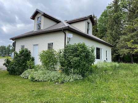 The exterior of the cottage is shown, featuring a light-colored siding and a unique roofline. Lush greenery surrounds the home, with bushes and plants adding a natural touch to the yard. The structure is set against a backdrop of trees and an overcast sky.