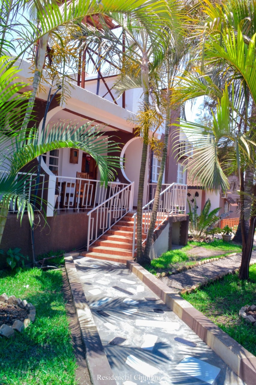 A well-maintained entrance path leads to a charming building, bordered by lush greenery and palm trees. A decorative railing enhances the walkway, which features a mix of stone and grass surfaces, inviting guests to enjoy the home's inviting exterior.