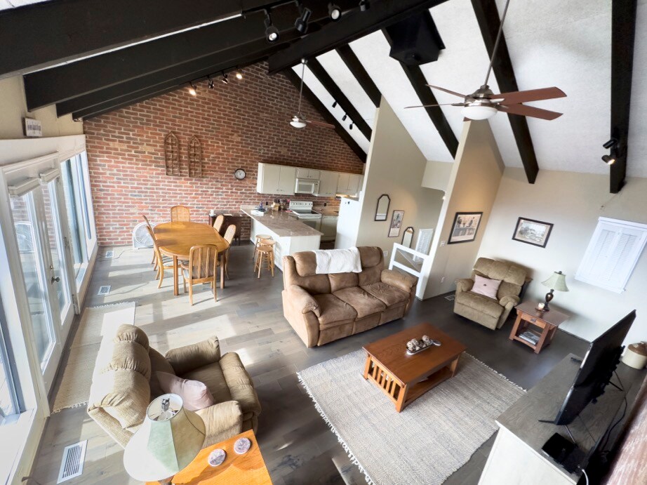 An open living area features a spacious seating arrangement with a comfortable sofa and armchairs. A dining table with wooden chairs is positioned nearby, and a modern kitchen is visible in the background. Exposed wooden beams and large windows bring in natural light.