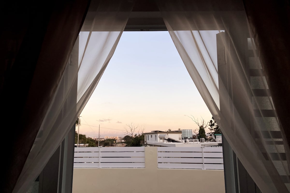 A serene view is framed by light, sheer curtains, showing a glimpse of an outdoor area. The sky displays soft pastel hues, indicating dusk. Fences and nearby buildings are visible, providing a sense of the neighborhood's tranquility.
