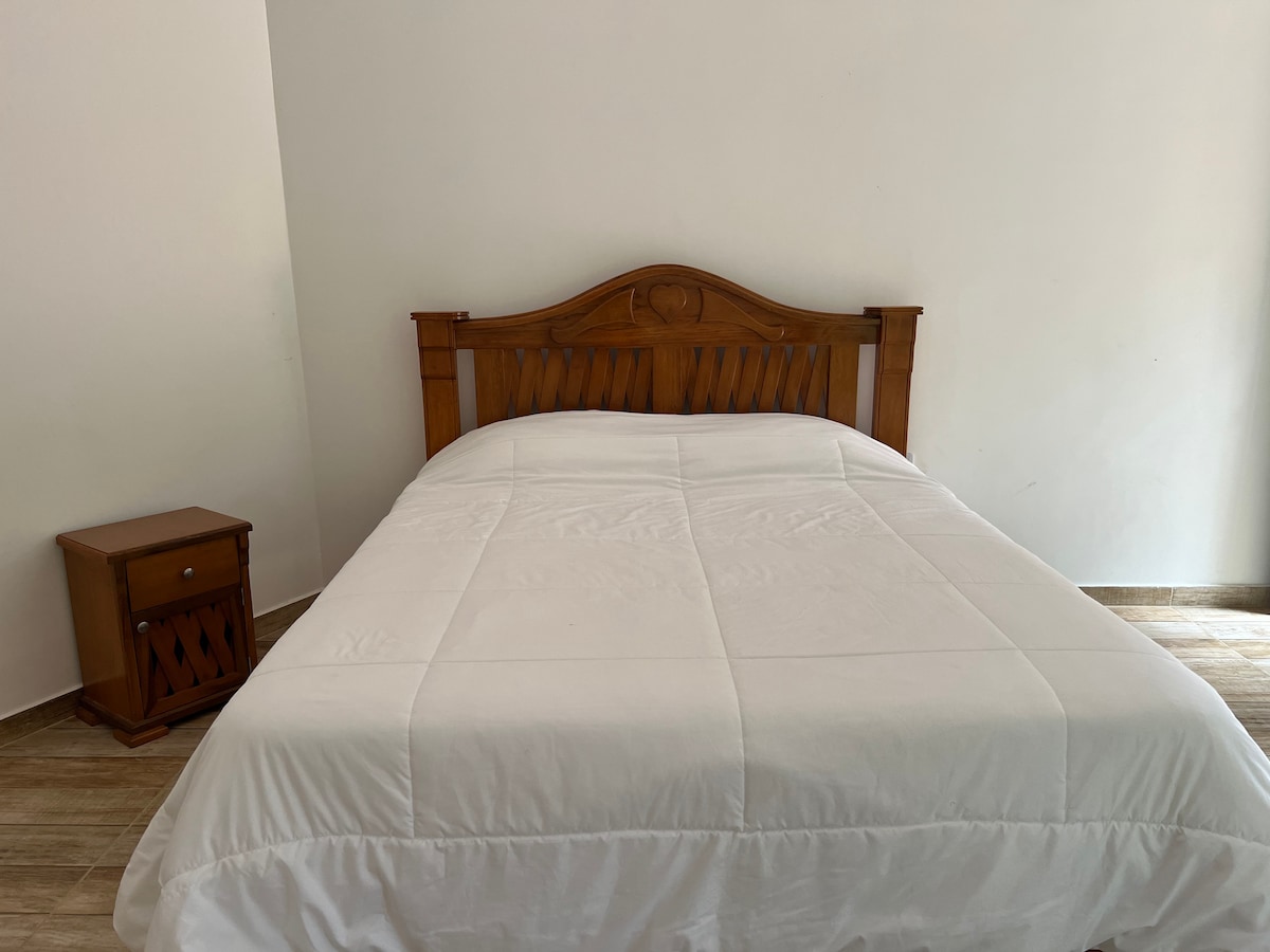 A spacious bed is centered against a white wall, featuring a wooden headboard. A neatly spread white comforter covers the mattress. Adjacent to the bed, a small wooden nightstand complements the overall simplicity of the room.