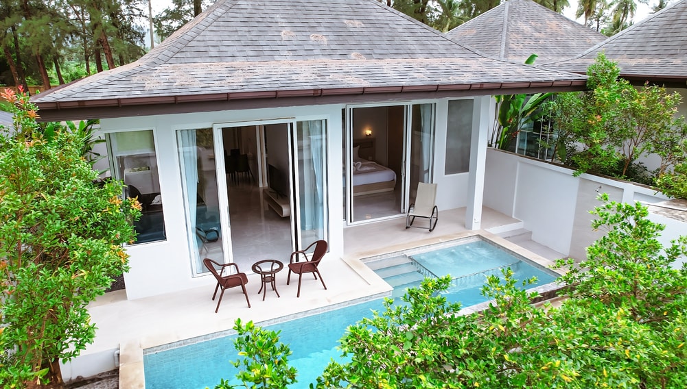 The exterior view showcases a modern building with a sleek design, featuring a private pool adjacent to a terrace. Comfortable seating options are provided, with a pair of chairs and a small table set in a green, landscaped area, surrounded by lush foliage.