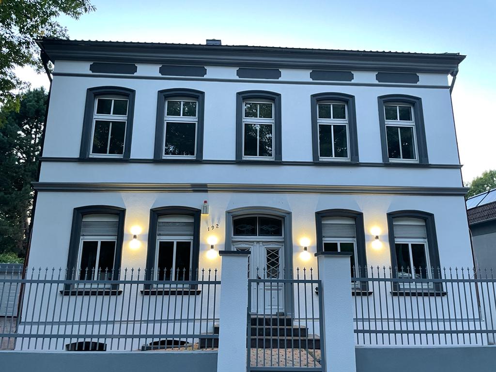 The exterior of the building is presented, featuring a classic architectural style with a crisp white facade. Several windows are symmetrically aligned on the upper and lower levels, framed in grey. Soft yellow lights illuminate the entrance and front fence, enhancing the inviting appearance.