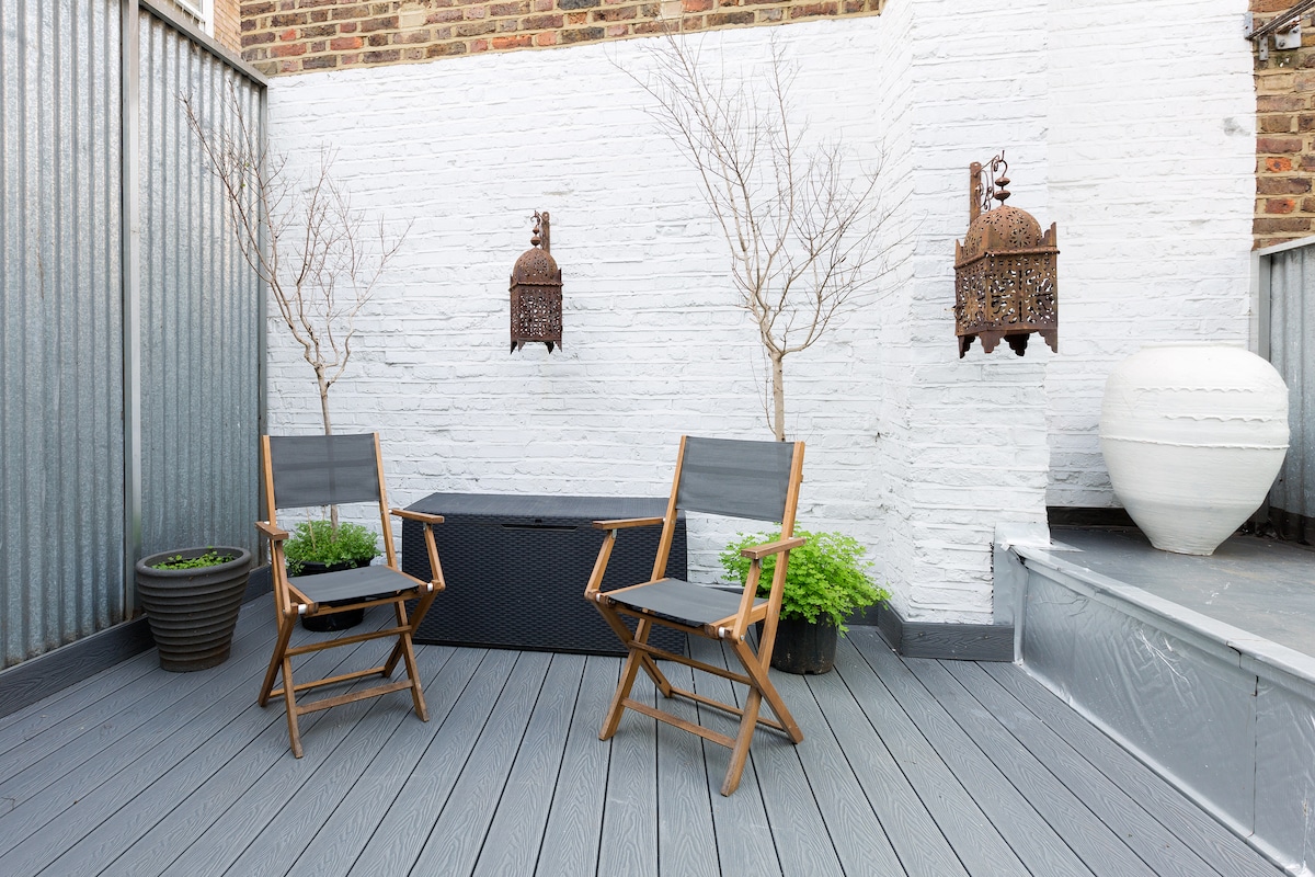 A cozy rooftop terrace features two wooden folding chairs positioned on a gray wooden deck. Potted plants bring greenery to the space, while decorative lanterns adorn the white brick wall, enhancing the outdoor ambiance.