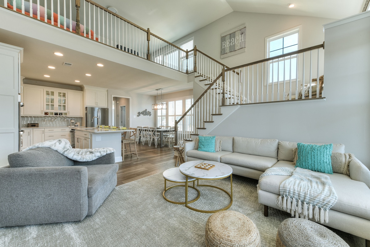 An open-concept living area reveals a light-filled space featuring a cozy sofa and an armchair. A pair of coffee tables sits on a plush rug, while a staircase leads to an upper level. The kitchen is visible, showcasing modern appliances and cabinetry.