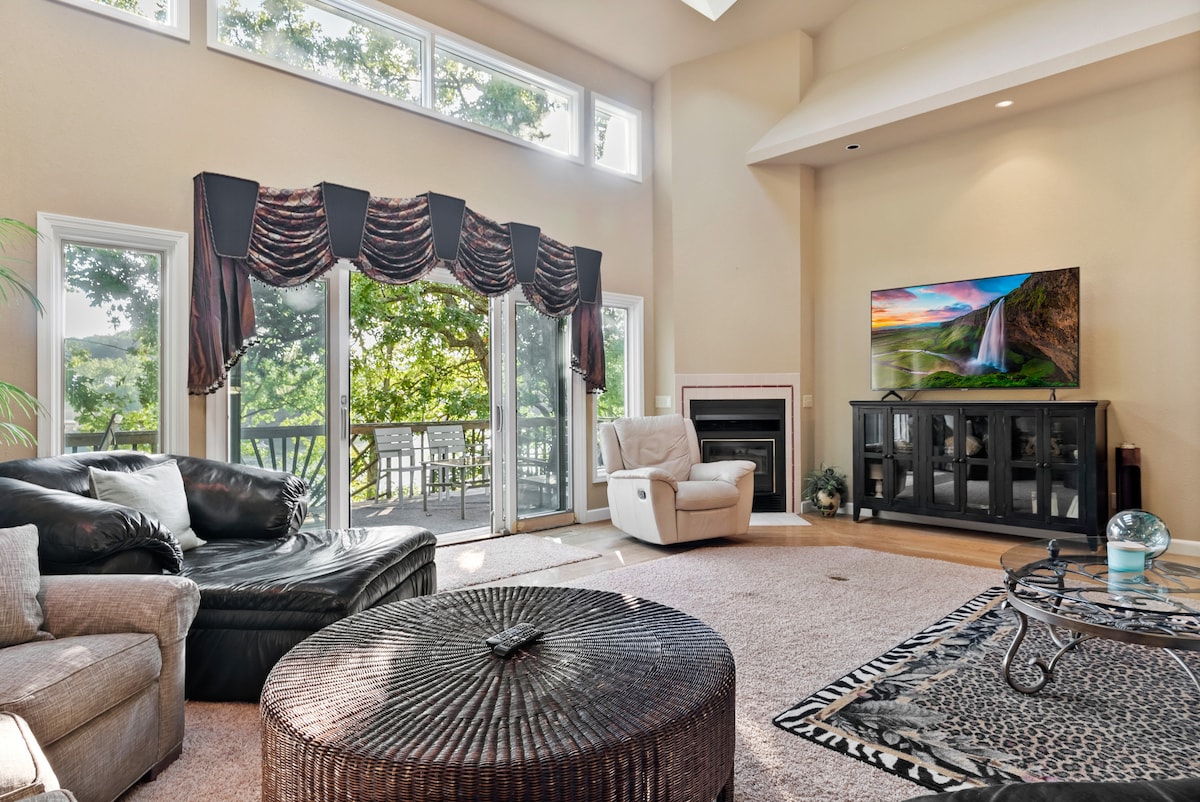 A bright living room is characterized by large windows that bring in natural light and offer views of greenery. A black leather sofa and a light-colored armchair provide seating options. A round, woven coffee table sits in the center, while an entertainment unit displays a television against a backdrop of a cozy fireplace.