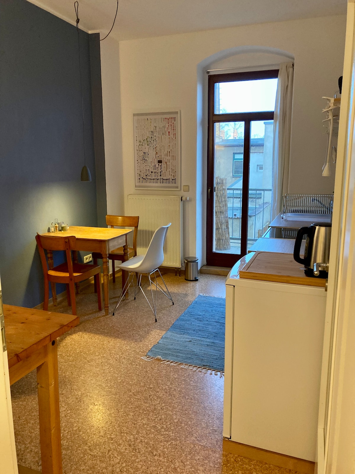 A cozy dining area features a wooden table surrounded by two chairs and a white chair. Natural light enters through a large window, illuminating the space. A small rug adds texture to the floor, while a kitchenette is visible at the side.