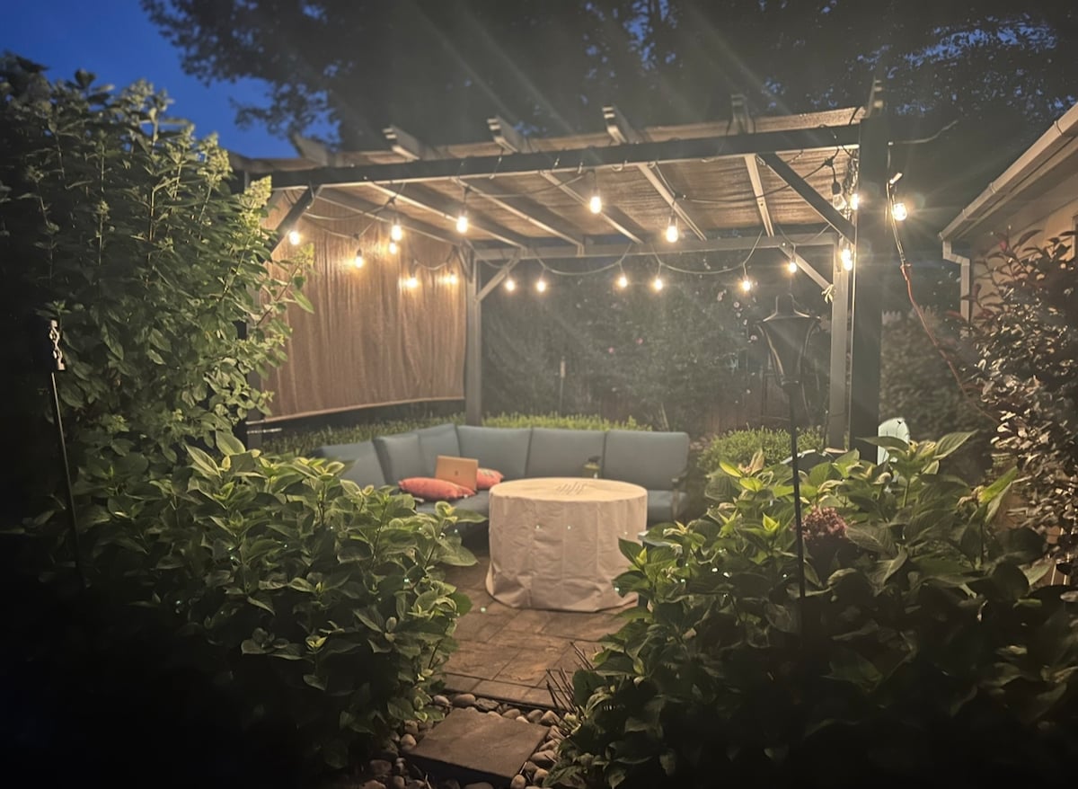 An inviting outdoor seating area is featured under a pergola adorned with warm string lights. The space includes a circular table surrounded by a sectional couch, creating a cozy spot for relaxation among lush greenery.